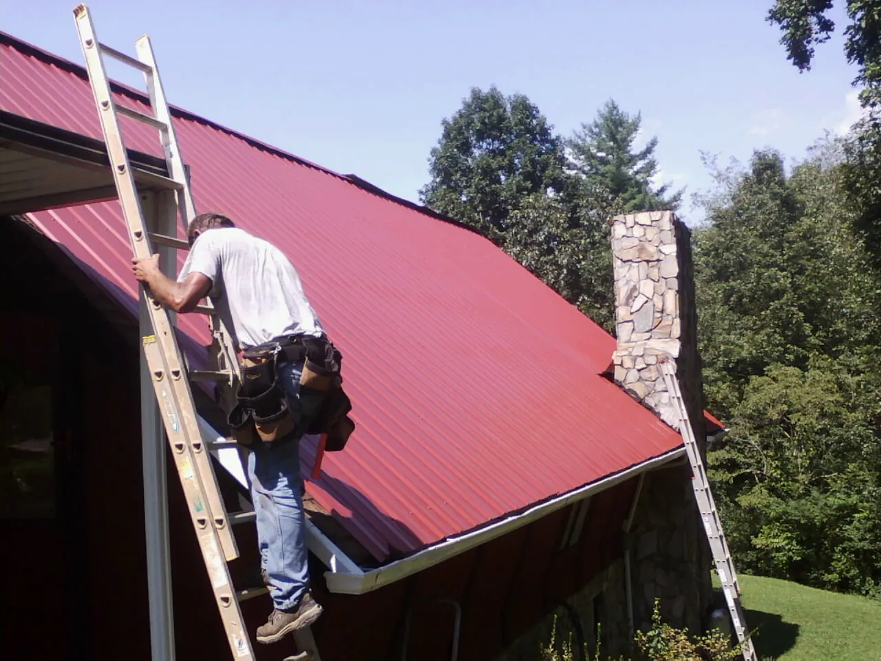 Roofer on a ladder beside a red metal roof and stone chimney — Soffit & Fascia Repair by Roofing Brothers in Orleans